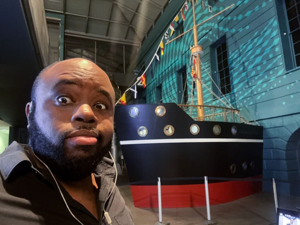 CEO of BridgeGap Media standing in front of a Caribbean-themed ship exhibit at the National Maritime Museum during the Windrush Food Culture event.